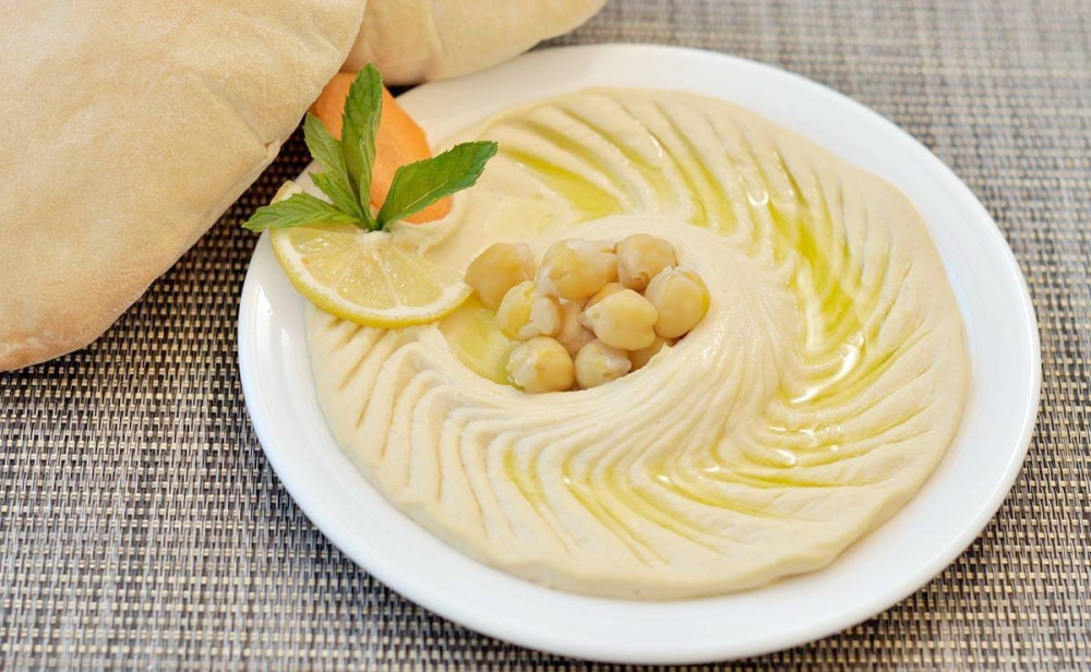 طبق حمص بالطحينة مزين بحبات الحمص الكاملة وزيت الزيتون مع خبز وخضار طازجة على طاولة خشبية